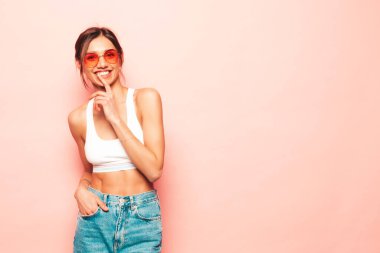 Beyaz formalı bluz ve kot pantolon giymiş güzel bir kadın. Seksi kaygısız neşeli model içeride eğleniyor. Stüdyoda pembe duvarın yanında poz veren sevimli ve pozitif kadın. Güneş gözlükleriyle