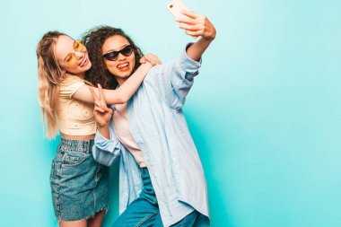 Moda kıyafetleri içinde iki güzel, gülümseyen uluslararası hippi kadın. Stüdyoda mavi duvarın yanında poz veren kaygısız seksi kadınlar. Pozitif modeller güneş gözlükleriyle eğleniyor. Selfie çekiyoruz.