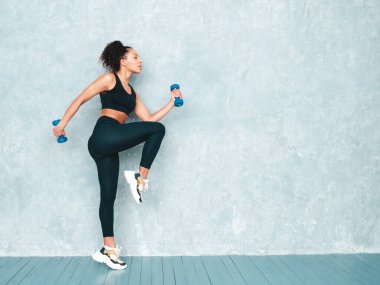 Spor elbiseli, saçları kıvırcık, spor elbiseli, gülümseyen bir kadın. Koşuyor, koşuyor. Mükemmel bronzlaşmış vücudu olan genç ve güzel bir manken. Stüdyoda gri duvarın yanında dambıllarla atlayan bir kadın.