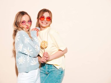 Moda kıyafetleri içinde gülümseyen iki güzel sarışın hippi kadın. Stüdyoda poz veren kaygısız seksi kadınlar. Moda ve pozitif modeller şekerleme lolipoplarıyla eğleniyorlar