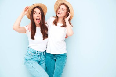 Son moda beyaz tişörtlü ve kot pantolonlu iki güzel gülümseyen hippi kadın stüdyoda açık mavi duvarın yanında poz veren kaygısız kadınlar. Şapkalı pozitif modeller. Mutlu ve neşeli.