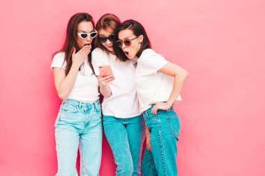 Üç güzel, gülümseyen hippi kadın, son moda tişört ve kot pantolon giyiyor. Stüdyoda pembe duvarın yanında poz veren kaygısız seksi kadınlar. Akıllı telefon ekranına bakan pozitif modeller. Uygulamalar kullanılıyor 