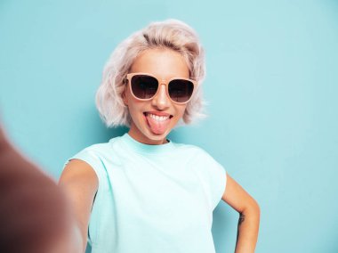 Moda kıyafetleri içinde gülümseyen güzel bir kadın. Stüdyoda mavi duvarın yanında poz veren kaygısız seksi kadın. Pozitif sarışın model güneş gözlükleriyle eğleniyor. Dilini gösteriyor. Pov selfie çekiliyor 