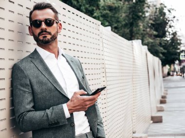Yakışıklı, kendine güvenen, stil sahibi, Lambersex modelinin portresi. Zarif takım elbise giymiş modern bir adam. Moda erkeği gün batımında çitin yanında poz veriyor. Güneş gözlükleriyle. Akıllı telefon uygulamaları kullanarak.