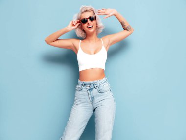 Moda kıyafetleri içinde gülümseyen güzel bir kadın. Stüdyoda mavi duvarın yanında poz veren kaygısız seksi kadın. Pozitif sarışın model eğleniyor ve deliriyor. Güneş gözlükleriyle