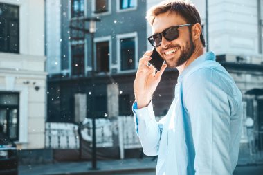 Yakışıklı, güler yüzlü, stil sahibi, lamberseksüel model. Mavi gömlekli, modern bir adam. Sokakta güneş gözlükleriyle poz veren modacı erkek. Gün batımında dışarıda. Akıllı telefon kullanarak. Konuşarak.