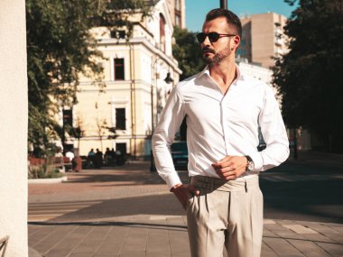 Yakışıklı, kendine güvenen, stil sahibi, lamberseksüel modelin portresi. Beyaz gömlek ve pantolon giymiş modern bir adam. Moda erkeği gün batımında Avrupa 'da sokak arkasındaymış gibi poz veriyor. Güneş gözlükleriyle