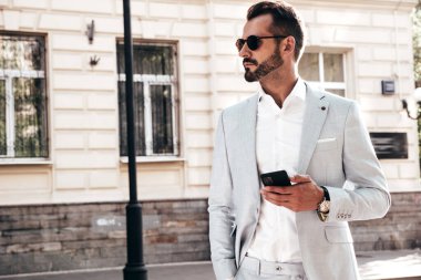 Yakışıklı, kendine güvenen, stil sahibi, Lambersex modelinin portresi. Zarif takım elbise giymiş modern bir adam. Moda erkeği gün batımında çitin yanında poz veriyor. Güneş gözlükleriyle. Akıllı telefon uygulamaları kullanarak.