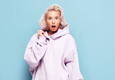 Moda kıyafetleri içinde gülümseyen güzel bir kadının portresi. Stüdyoda mavi duvarın yanında poz veren kaygısız seksi kadın. Pozitif sarışın model eğleniyor ve deliriyor. Şok oldum ve şaşırdım.