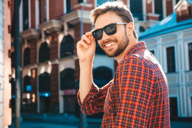 Yakışıklı, şık, havalı, havalı, lamberseksüel model. Kırmızı kareli gömlek giymiş modern bir adam. Moda erkeği güneş gözlükleriyle gökdelenin yanında poz veriyor. Gün batımında dışarıda. 