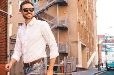 Yakışıklı, gülümseyen stil sahibi Lambersex modelinin portresi. Beyaz gömlek giymiş modern bir adam. Sokakta güneş gözlükleriyle poz veren modacı erkek. Gün batımında dışarıda. 