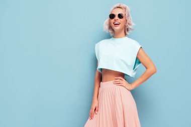 Moda kıyafetleri içinde gülümseyen güzel bir kadın. Stüdyoda mavi duvarın yanında poz veren kaygısız seksi kadın. Pozitif sarışın model eğleniyor ve deliriyor. Güneş gözlükleriyle