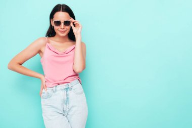 Moda kıyafetleri içinde gülümseyen güzel bir kadının portresi. Stüdyoda mavi duvarın yanında poz veren kaygısız seksi kadın. Pozitif model içeride eğleniyor. Neşeli ve mutlu. Güneş gözlükleriyle
