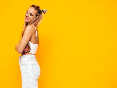 Moda kıyafetleri içinde gülümseyen sarışın güzel bir kadının portresi. Stüdyoda sarı duvarın yanında poz veren kaygısız seksi kadın. Pozitif model içeride eğleniyor. Neşeli ve mutlu