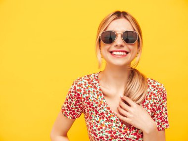 Moda kıyafetleri içinde gülümseyen sarışın güzel bir kadının portresi. Stüdyoda sarı duvarın yanında poz veren kaygısız bir kadın. Pozitif model içeride eğleniyor. Neşeli ve mutlu. Güneş gözlükleriyle