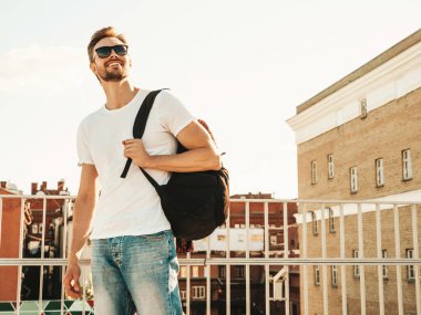 Yakışıklı, güler yüzlü, Lambersex mankeni. Beyaz tişört ve kot giyen şık bir adam. Moda erkek plajda çitlerin yanında güneş gözlüklü okul çantasıyla poz veriyor.