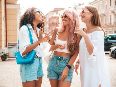 Son moda yaz kıyafetleri içinde gülümseyen üç genç bayan. Sokakta poz veren kaygısız, çok ırklı kadınlar. Güneş gözlükleriyle eğlenen pozitif modeller. Neşeli ve mutlu