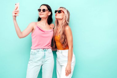 Moda kıyafetleri içinde gülümseyen iki güzel esmer hippi kadının portresi. Mavi duvarın yanında poz veren kaygısız seksi kadınlar. Pozitif modeller eğleniyor. Neşeli ve mutlu. selfie çekmek 
