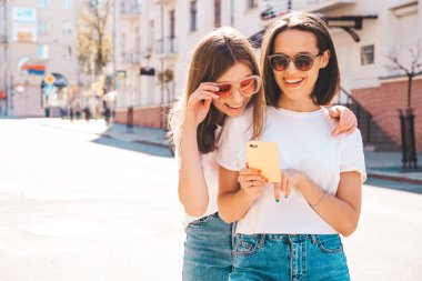 Son moda beyaz tişörtlü ve kot pantolonlu iki güzel gülümseyen yeniyetme kadın. Sokakta arka planda poz veren kaygısız kadınlar. Akıllı telefon uygulamaları kullanan, ekrana bakan pozitif modeller.