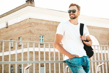 Yakışıklı, güler yüzlü, Lambersex mankeni. Beyaz tişört ve kot giyen şık bir adam. Moda erkek plajda çitlerin yanında güneş gözlüklü okul çantasıyla poz veriyor.