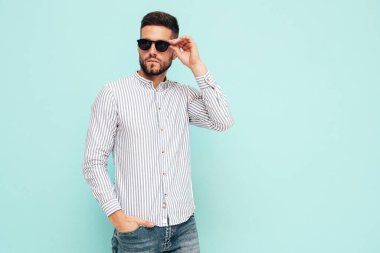 Yakışıklı, kendinden emin bir modelin portresi. Gömlek ve kot giyen seksi şık bir adam. Moda hippi erkek stüdyoda mavi duvarın yanında poz veriyor. Güneş gözlükleriyle