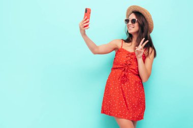 Son moda kırmızı elbisesi içinde gülümseyen güzel bir kadın. Stüdyoda mavi duvarın yanında poz veren kaygısız seksi kadın. Pozitif esmer model şapka ve güneş gözlükleriyle eğleniyor. Selfie çekiyoruz.