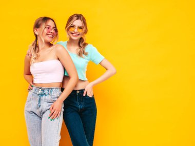 Moda kıyafetleri içinde gülümseyen iki güzel sarışın hippi kadın. Stüdyoda sarı duvarın yanında poz veren kaygısız seksi kadınlar. Pozitif modeller eğleniyor. Neşeli ve mutlu. Güneş gözlükleriyle