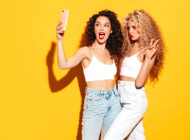 Kırmızı yaz mayo giymiş, gülümseyen iki güzel hippi kadın. Saç stili kıvırcık saçlı moda modeller stüdyoda eğleniyor. Sarıda izole edilmiş seksi bir kadın. Selfie çekiyoruz.