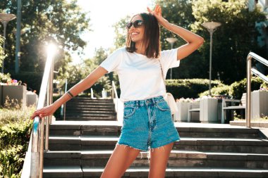 Güzel gülümseyen hippi bir kadının son moda kot şortlu portresi. Günbatımında sokakta poz veren kaygısız seksi manken. Güneş gözlüklü pozitif model.