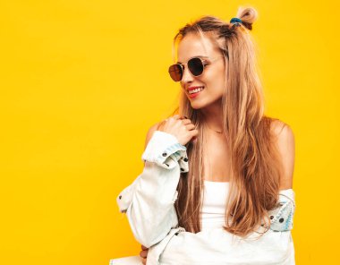 Moda kıyafetleri içinde gülümseyen sarışın güzel bir kadının portresi. Stüdyoda sarı duvarın yanında poz veren kaygısız seksi kadın. Pozitif model içeride eğleniyor. Neşeli ve mutlu güneş gözlükleriyle