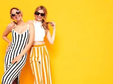 Moda kıyafetleri içinde gülümseyen iki güzel sarışın hippi kadın. Stüdyoda sarı duvarın yanında poz veren kaygısız seksi kadınlar. Pozitif modeller eğleniyor. Neşeli ve mutlu. Güneş gözlükleriyle