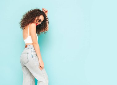 Yeni moda hippi kıyafetleri içinde gülümseyen güzel bir kadın. Stüdyoda mavi duvarın yanında poz veren kaygısız bir kadın. Kıvırcık saçlı esmer bir model. Neşeli ve mutlu. Güneş gözlükleriyle