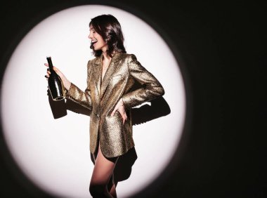 Parlak makyajlı, elinde şampanya şişesi olan, beyaz duvarın yanında poz veren kaygısız bir kadın.