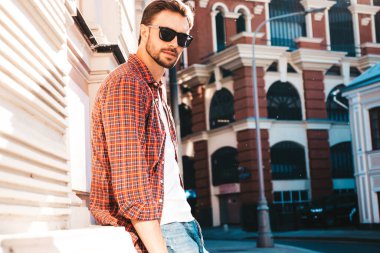 Yakışıklı, kendine güvenen, havalı, havalı, lamberseksüel model. Kırmızı kareli gömlek giymiş modern bir adam. Moda erkeği güneş gözlükleriyle gökdelenin yanında poz veriyor. Gün batımında dışarıda. 