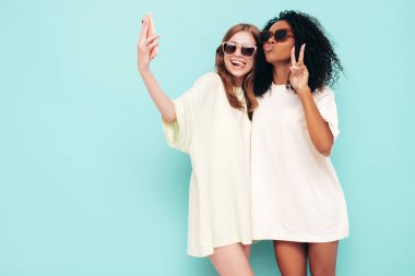 Moda kıyafetleri içinde iki güzel, gülümseyen uluslararası hippi kadın. Stüdyoda mavi duvarın yanında poz veren kaygısız seksi kadınlar. Pozitif modeller güneş gözlükleriyle eğleniyor. Selfie çekiyoruz.