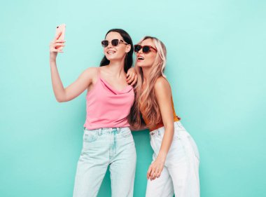 Moda kıyafetleri içinde gülümseyen iki güzel esmer hippi kadının portresi. Mavi duvarın yanında poz veren kaygısız seksi kadınlar. Pozitif modeller eğleniyor. Neşeli ve mutlu. selfie çekmek 