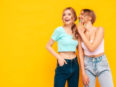 İki güzel, gülümseyen esmer hippi kadın moda yaz kıyafetleri içinde. Stüdyoda sarı duvarın yanında poz veren kaygısız seksi kadınlar. Pozitif model arkadaşına sırrını, dedikodusunu söyler.