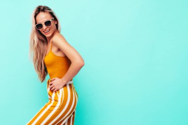 Son moda sarı elbiseli, gülümseyen, güzel sarışın kadın. Stüdyoda mavi duvarın yanında poz veren kaygısız seksi kadın. Pozitif model içeride eğleniyor. Neşeli ve mutlu. Güneş gözlükleriyle 