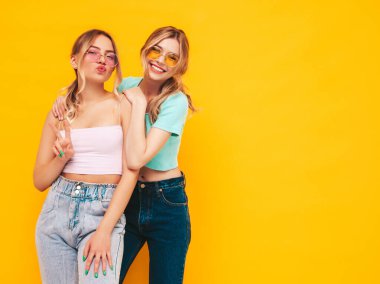 Moda kıyafetleri içinde gülümseyen iki güzel sarışın hippi kadın. Stüdyoda sarı duvarın yanında poz veren kaygısız seksi kadınlar. Pozitif modeller eğleniyor. Neşeli ve mutlu. Güneş gözlükleriyle