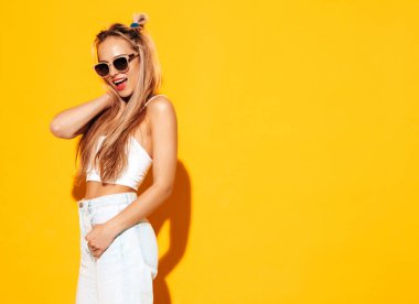 Moda kıyafetleri içinde gülümseyen sarışın güzel bir kadının portresi. Stüdyoda sarı duvarın yanında poz veren kaygısız seksi kadın. Pozitif model içeride eğleniyor. Neşeli ve mutlu güneş gözlükleriyle