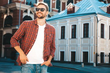 Yakışıklı, şık, havalı, havalı, lamberseksüel model. Kırmızı kareli gömlek giymiş modern bir adam. Moda erkeği güneş gözlükleriyle gökdelenin yanında poz veriyor. Gün batımında dışarıda. 