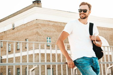 Yakışıklı, gülümseyen hipster Lambersex modelinin portresi. Beyaz tişört ve kot giyen şık bir adam. Moda erkeği sahildeki çitlerin yanında sokak arkasında poz veriyor. Sırt çantasıyla  