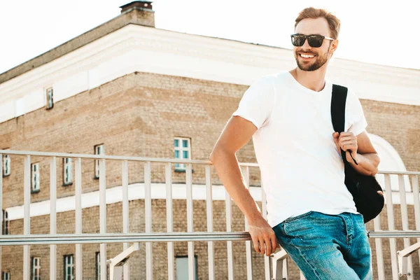 Yakışıklı, gülümseyen hipster Lambersex modelinin portresi. Beyaz tişört ve kot giyen şık bir adam. Moda erkeği sahildeki çitlerin yanında sokak arkasında poz veriyor. Sırt çantasıyla  