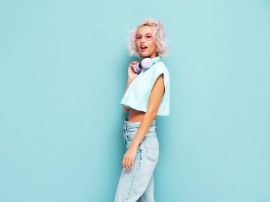 Moda kıyafetleri içinde gülümseyen güzel bir kadın. Stüdyoda mavi duvarın yanında poz veren kaygısız seksi kadın. Pozitif sarışın model eğleniyor. Kablosuz kulaklıkla müzik dinlemek