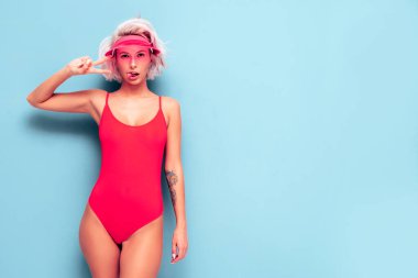Yazın mayo giymiş gülümseyen sarışın modelin portresi kırmızı mayo ve şeffaf şapka. Seksi kaygısız kadın eğleniyor ve deliriyor. Stüdyoda mavi duvarın yanında poz veren kadın.