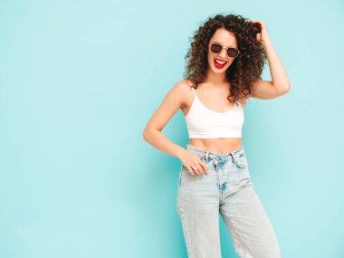 Yeni moda hippi kıyafetleri içinde gülümseyen güzel bir kadın. Stüdyoda mavi duvarın yanında poz veren kaygısız bir kadın. Kıvırcık saçlı esmer bir model. Neşeli ve mutlu. Güneş gözlükleriyle