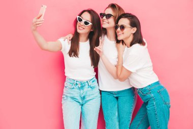 Aynı yaz moda olan beyaz tişört ve kot pantolon giyen üç güzel, gülümseyen hippi kadın. Stüdyoda pembe duvarın yanında poz veren kaygısız seksi kadınlar. Pozitif modeller selfie çekiyor.