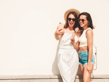 Son moda yaz kıyafetleri içinde iki güzel, gülümseyen hippi kadın. Sokakta beyaz şapkalı, kaygısız kadınlar poz veriyor. Pozitif saf modeller gün batımında eğleniyorlar, selfie çekiyorlar.