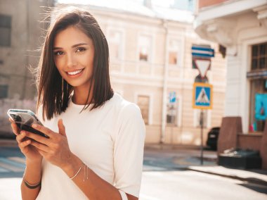 Modayı takip eden yaz elbisesi giymiş güzel, gülümseyen hippi kadın. Günbatımında sokakta poz veren kaygısız seksi kadın. Pozitif model. Akıllı telefon uygulamaları kullanarak. Cep telefonu ekranına bakıyorum.