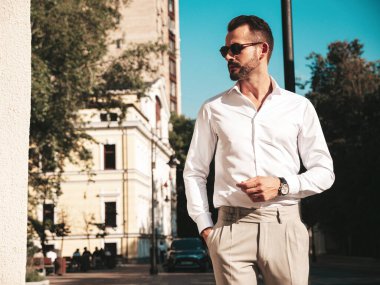 Yakışıklı, kendine güvenen, stil sahibi, lamberseksüel modelin portresi. Beyaz gömlek ve pantolon giymiş modern bir adam. Moda erkeği gün batımında Avrupa 'da sokak arkasındaymış gibi poz veriyor. Güneş gözlükleriyle